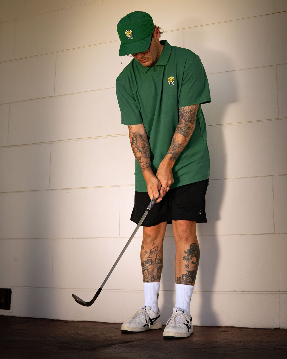 A man wearing a green polo shirt and matching cap swings a golf club while standing against a wall.