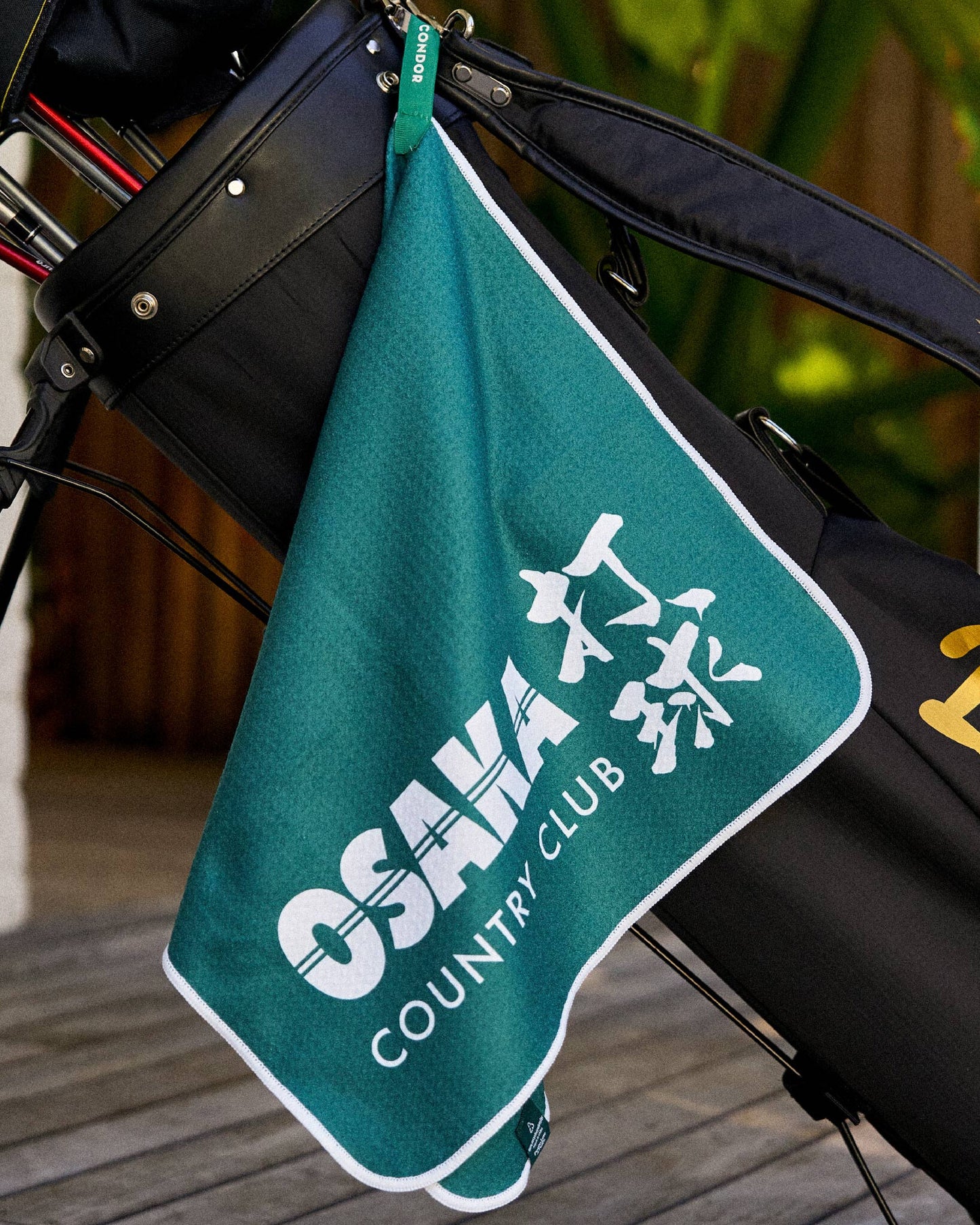 Alpine green golf towel hanging from a black golf bag, featuring a white Osaka Country Club graphic print.