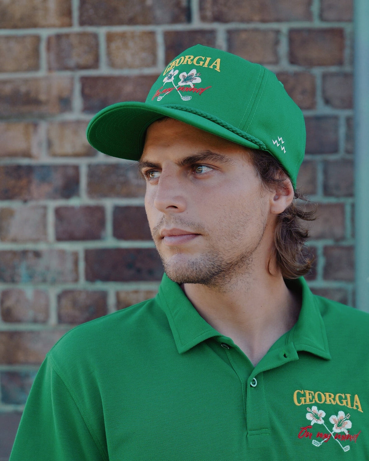 A man wearing a green snapback hat and matching polo shirt, both featuring embroidered flower designs.