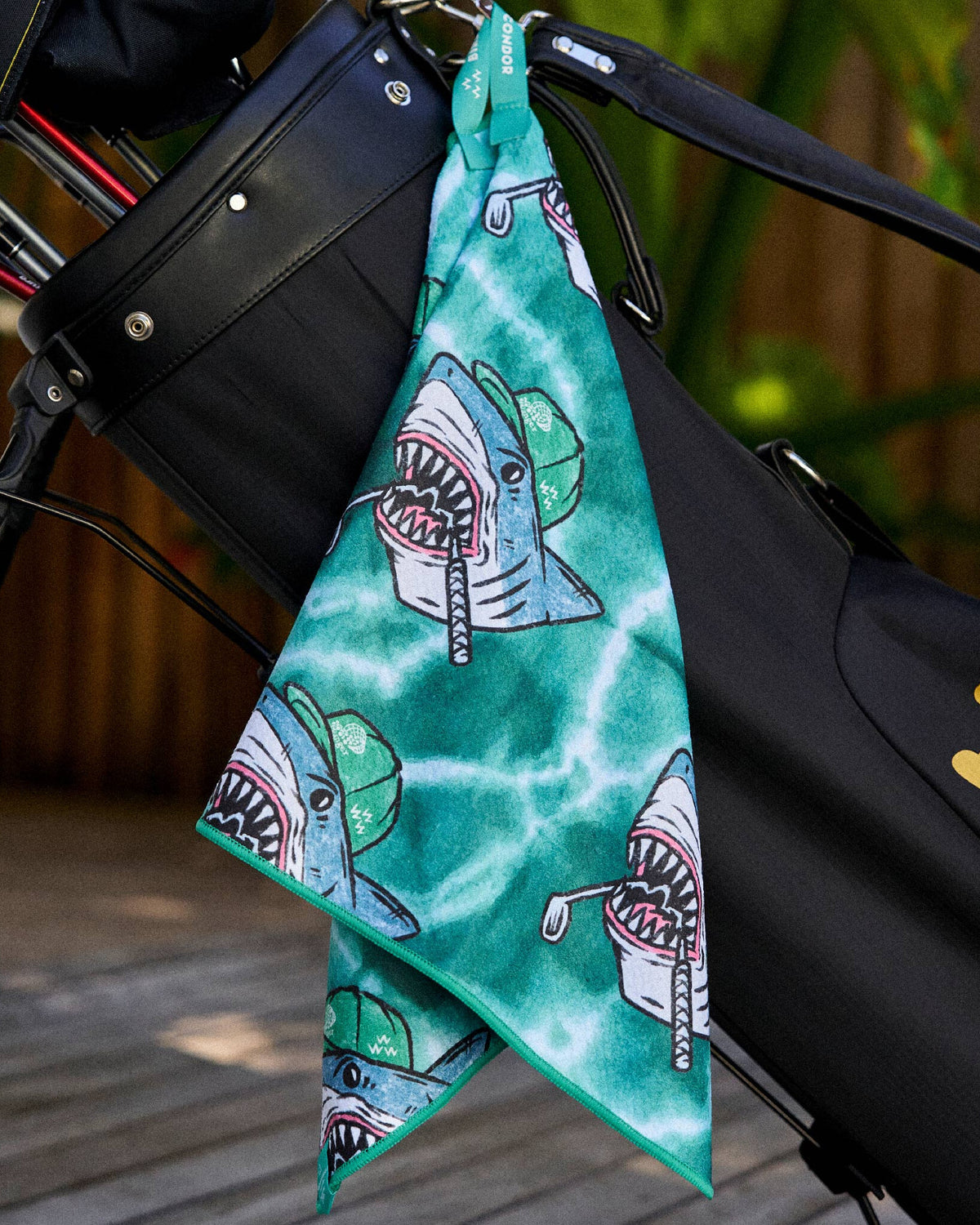 Green tie-dye golf towel hanging from a black golf bag, printed with sharks wearing golf caps.