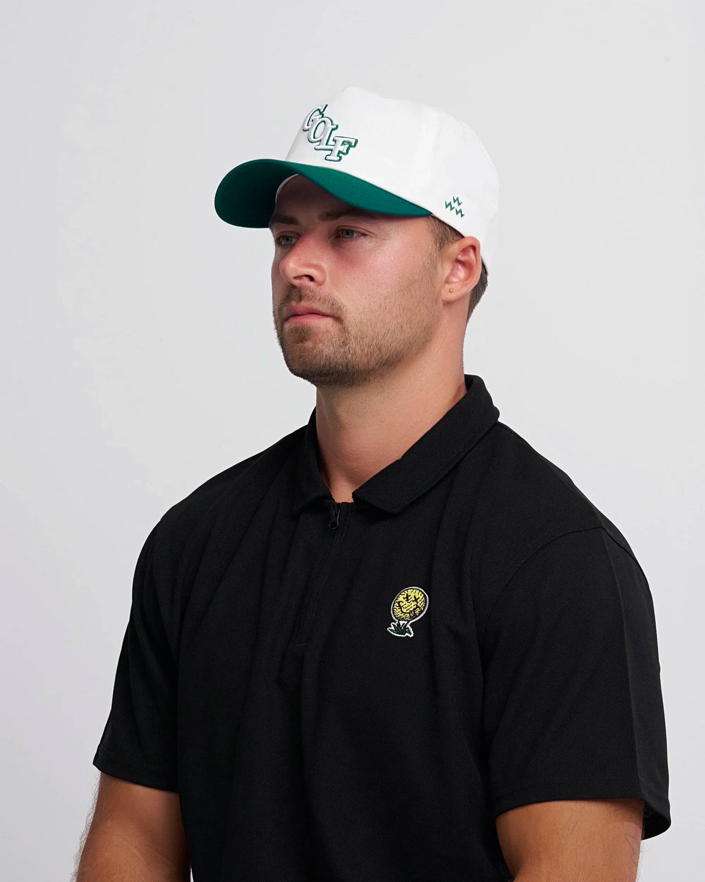 Man wearing a black polo shirt with a neverfind yellow golf ball logo and a white cap with green brim and logo on a gray background