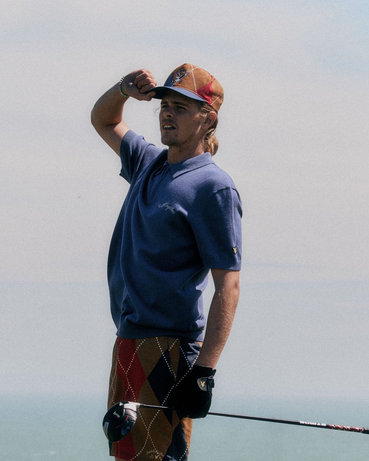 A man wearing a crown blue polo shirt, argyle print shorts, and a matching patterned cap outdoors.