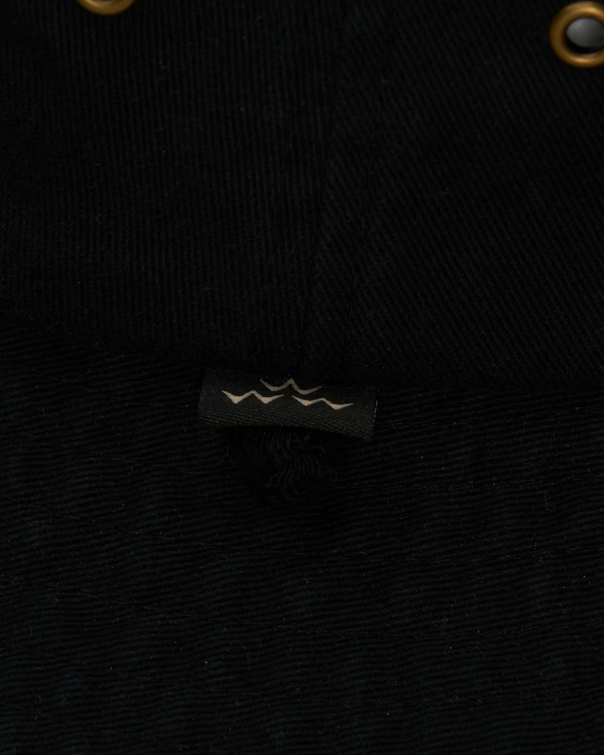 Detail of the black boonie hat showing brass eyelets and a small woven label featuring a wave design.