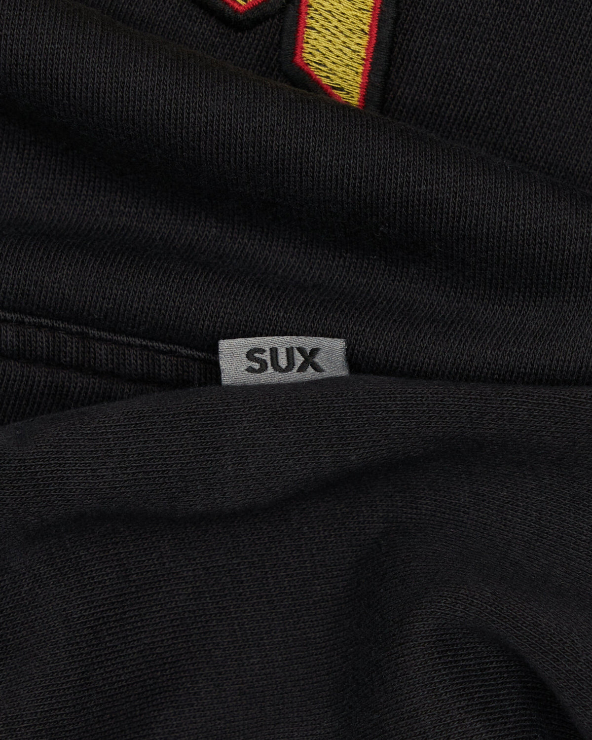 Detail of a black cotton hoodie featuring a small grey woven brand tag with SUX text.