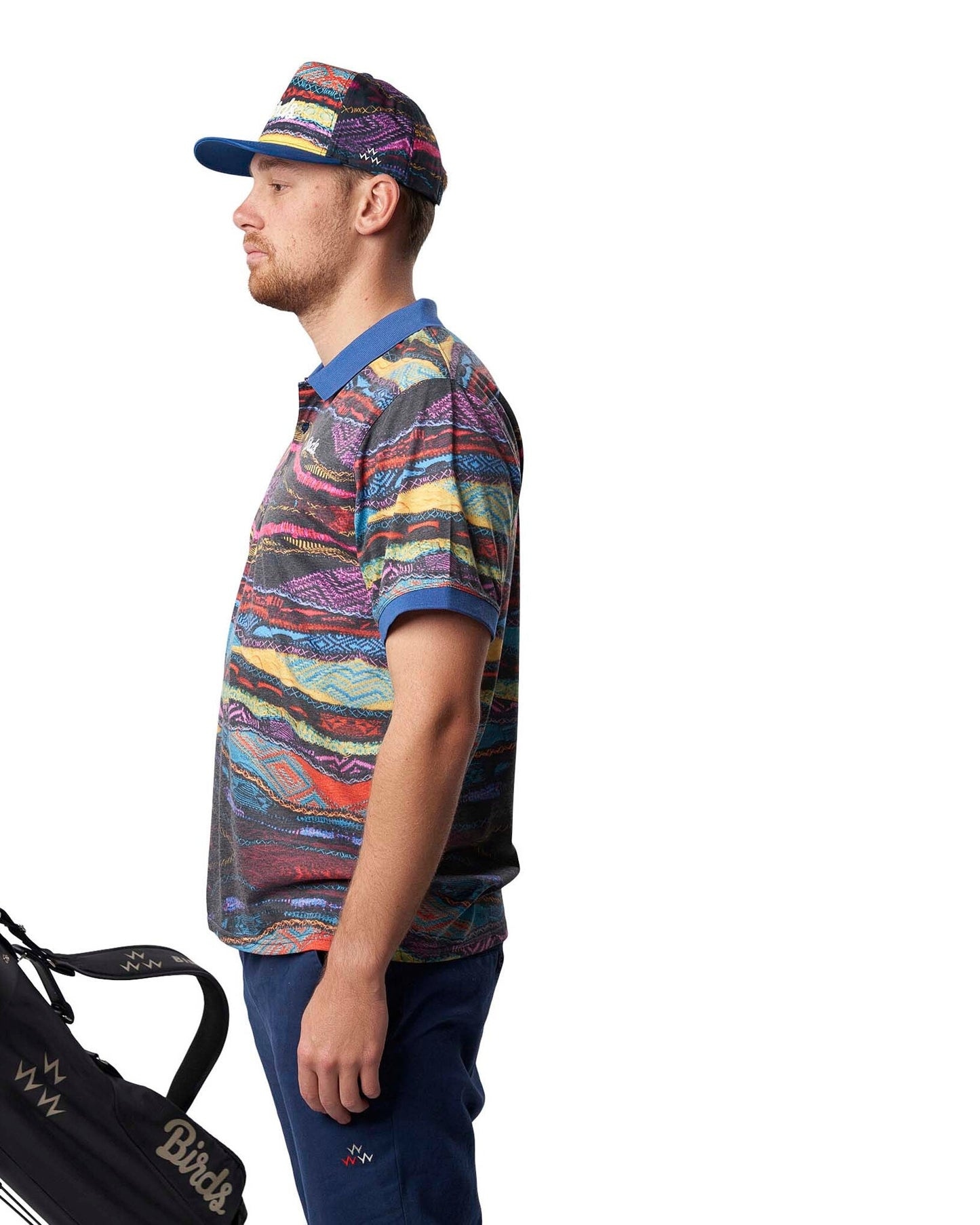 A man wearing a Coogi print cap and matching polo shirt beside a golf bag.