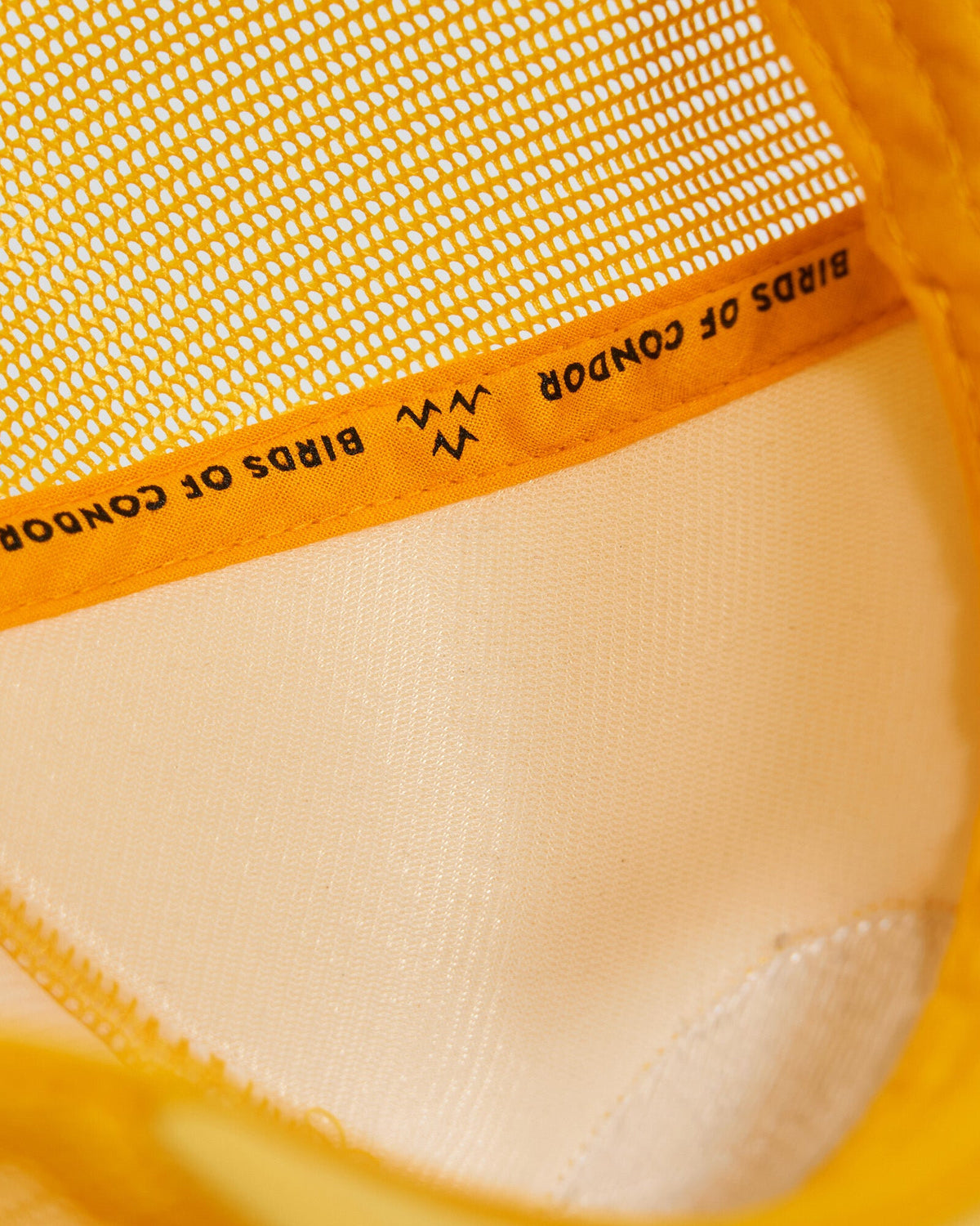 Detail of a yellow trucker cap showing internal orange taping with black branded text and mesh lining.