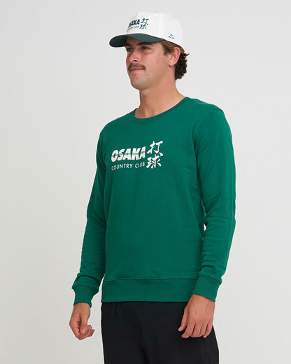 Man wearing a green sweatshirt with 'Osaka Country Club' logo by Birds of Condor and a white cap on a gray background