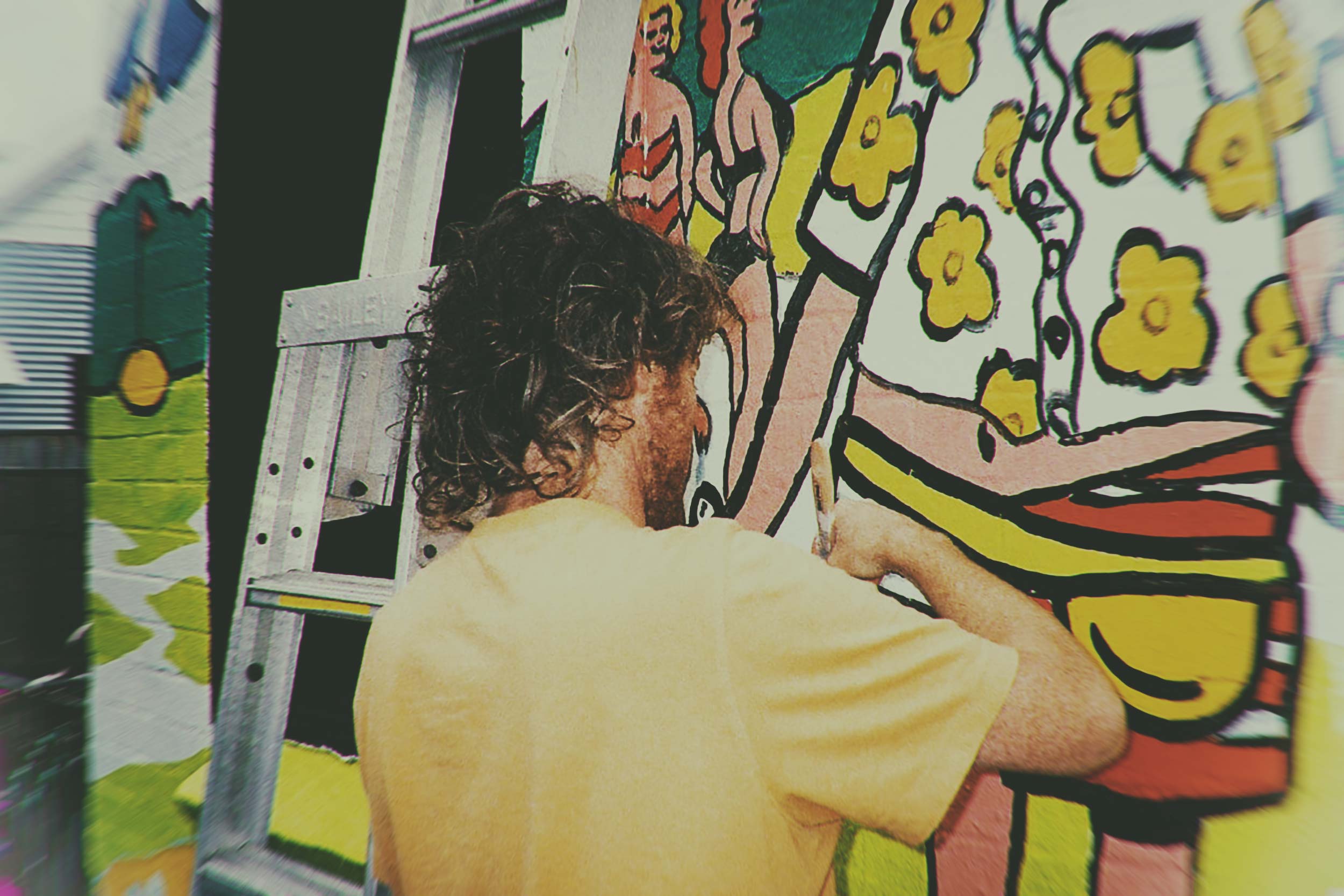 A man in a yellow t-shirt painting a colorful mural on a ladder, with a stylized cartoon figure and yellow flowers visible.