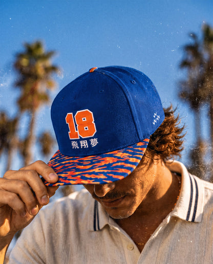 A man wearing a blue snapback cap with orange number 18 and tiger camo brim. He is holding the brim in front of his eyes. The background shows a sunny blue sky with blurred palm trees.