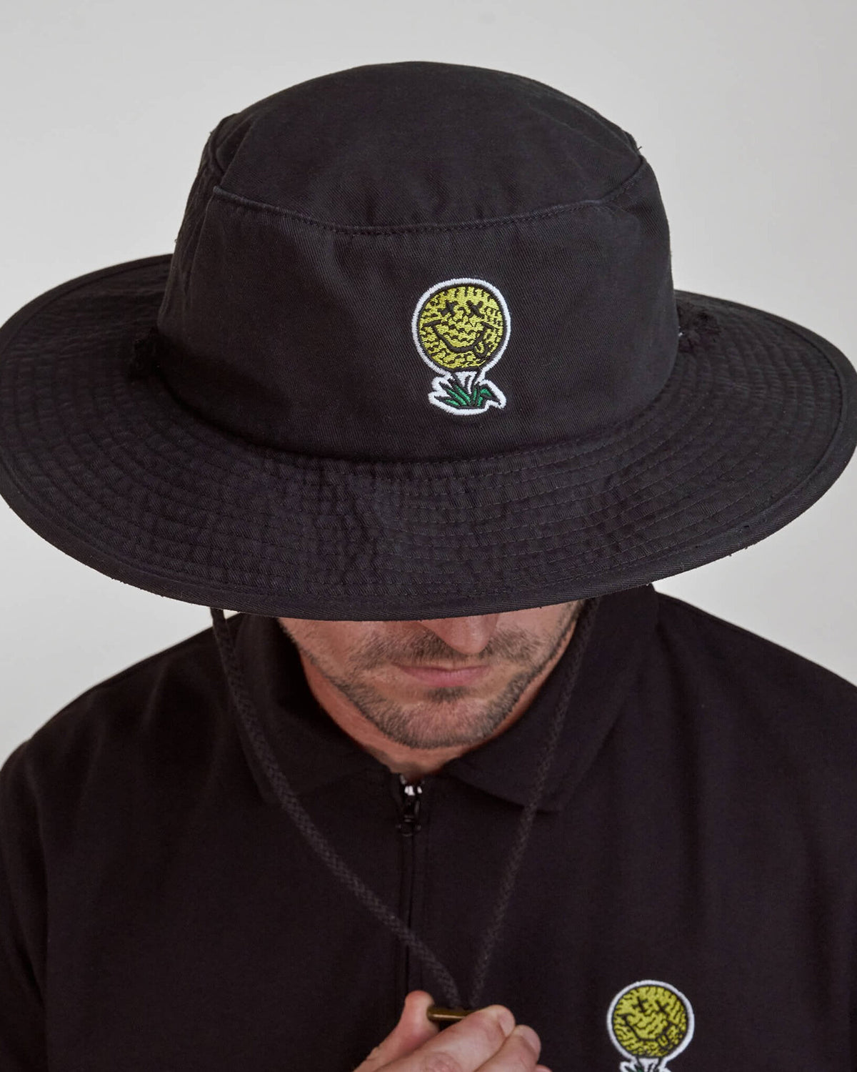 A person wearing a black boonie hat featuring a yellow and white smiley face golf ball logo design.