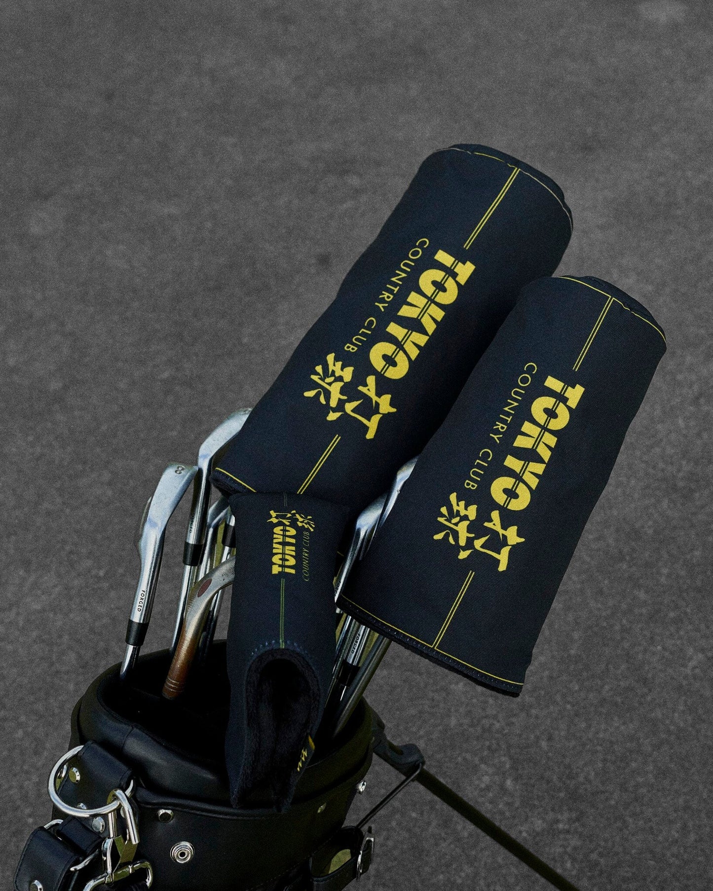 Detail of black headcovers with yellow lettering and piping, placed inside a golf bag on asphalt.
