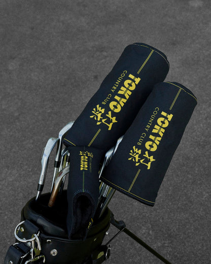 Detail of black headcovers with yellow lettering and piping, placed inside a golf bag on asphalt.