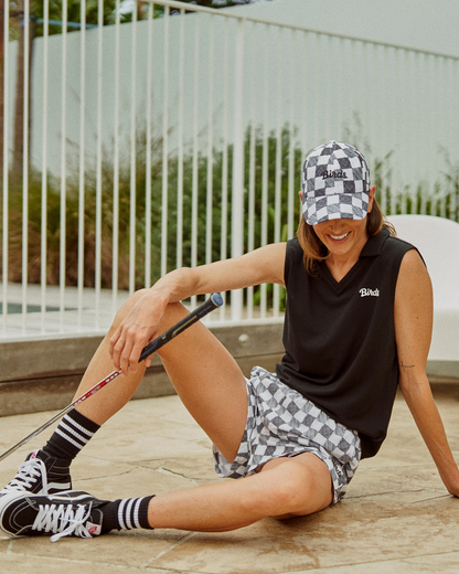 A woman wearing a black and white gingham cap, black polo top, and matching shorts, holding a golf club.