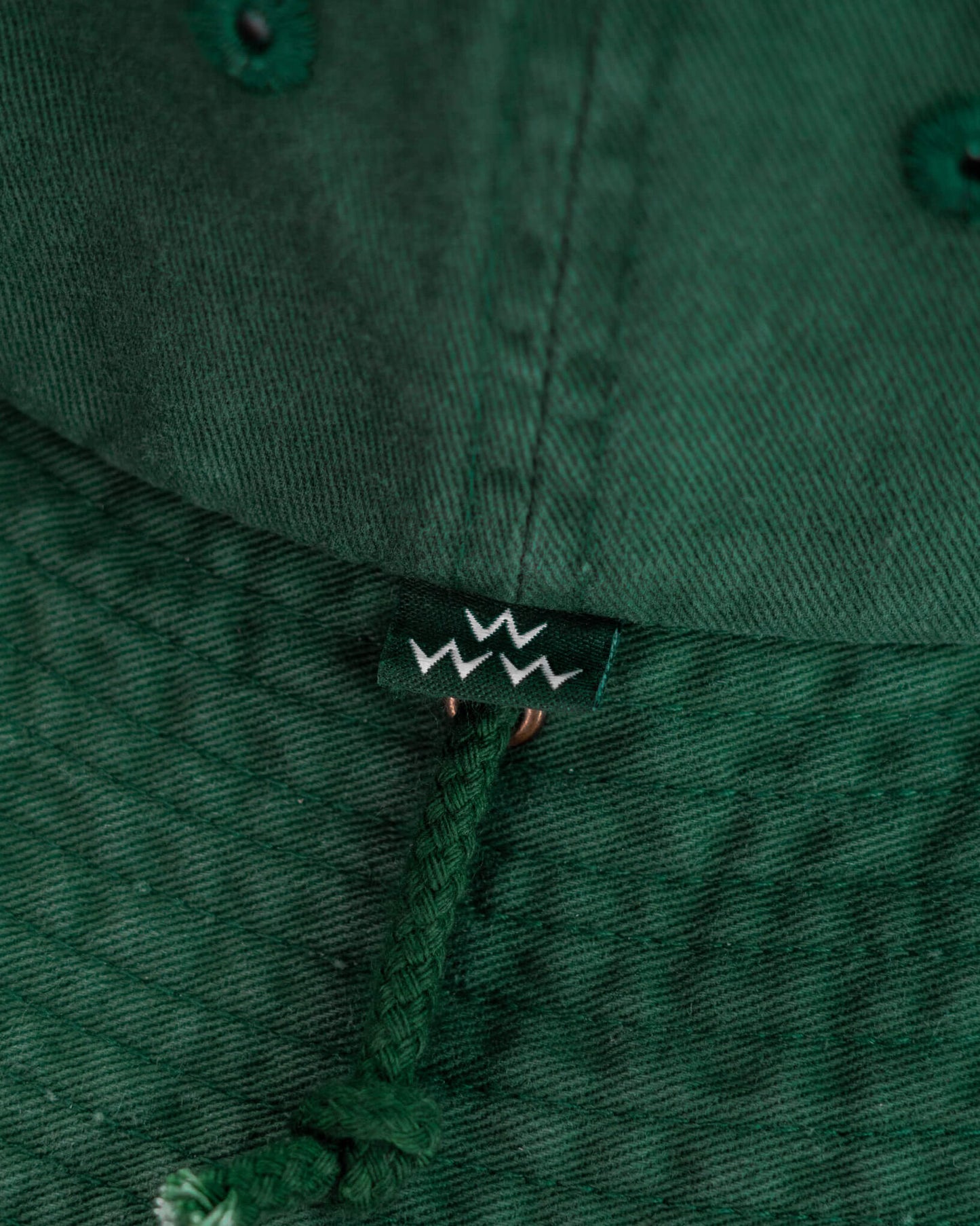 Detail of a green boonie hat featuring a small branded tag and an attached braided chin cord.