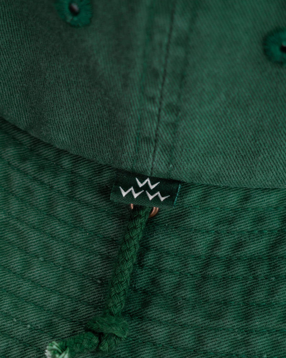 Detail of a green boonie hat featuring a small branded tag and an attached braided chin cord.