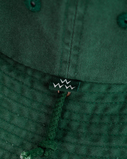 Detail of a green boonie hat featuring a small branded tag and an attached braided chin cord.