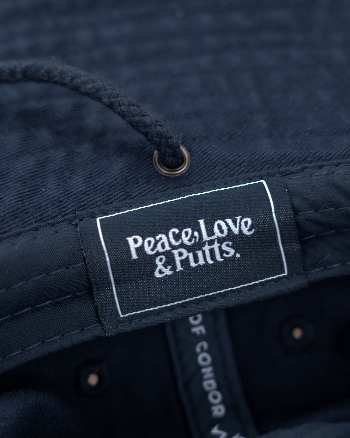 Detail of a navy boonie hat showing a woven label with white Peace, Love & Putts. text.