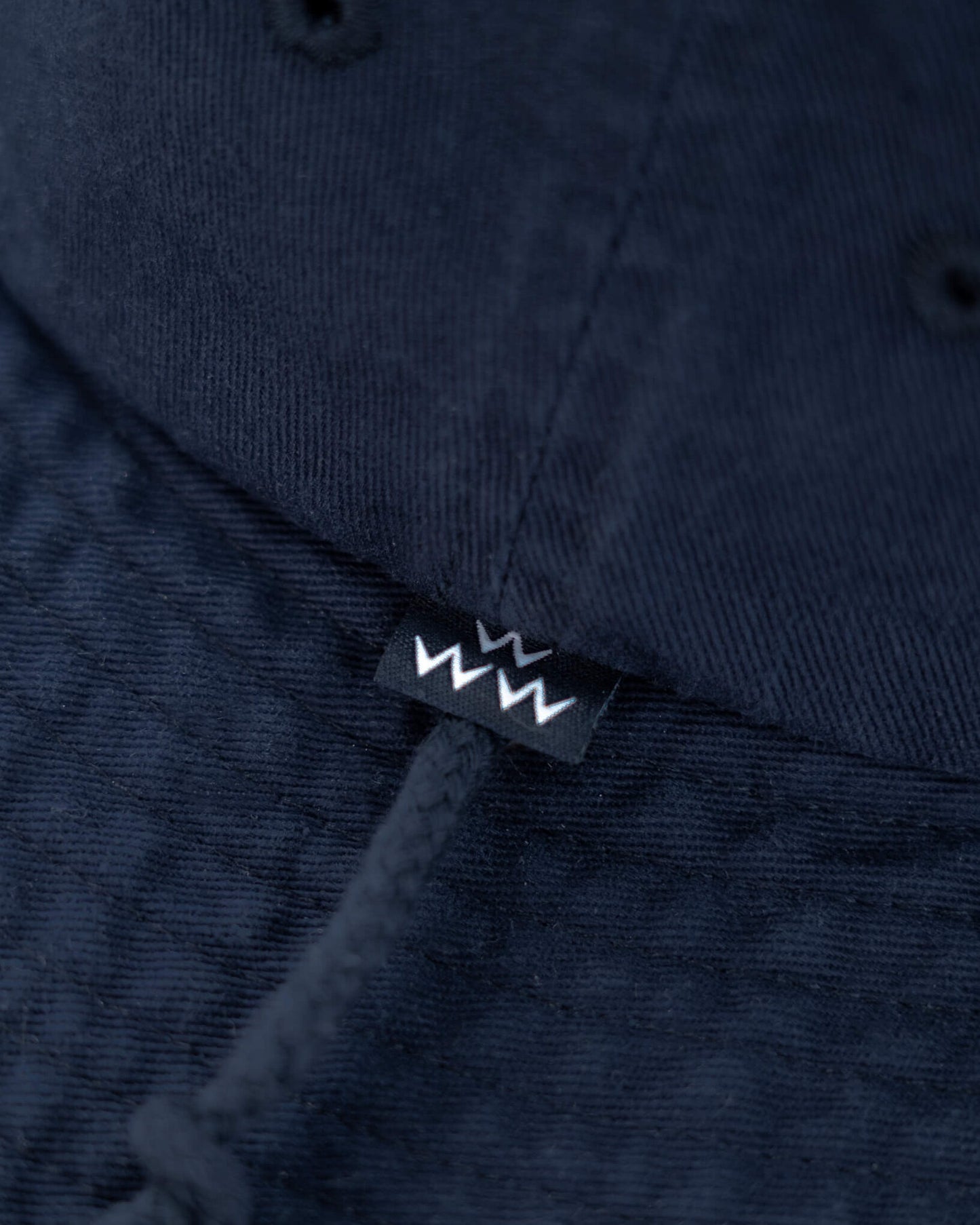 Detail of a navy boone hat featuring a black woven brand label with white zig-zag graphic and chin cord.