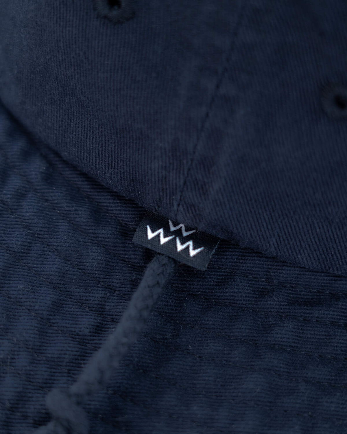 Detail of a navy boone hat featuring a black woven brand label with white zig-zag graphic and chin cord.