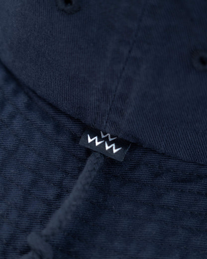 Detail of a navy boone hat featuring a black woven brand label with white zig-zag graphic and chin cord.