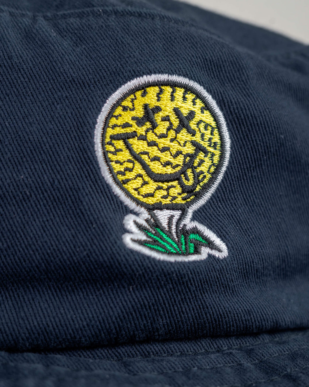 Detail of a navy cotton boonie featuring a yellow smiley face golf ball logo with X eyes on a white tee.
