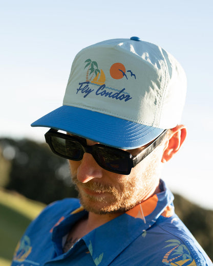 A man wearing a sky blue five-panel nylon snapback cap with a sunset, palm tree, and Fly Condor graphic.