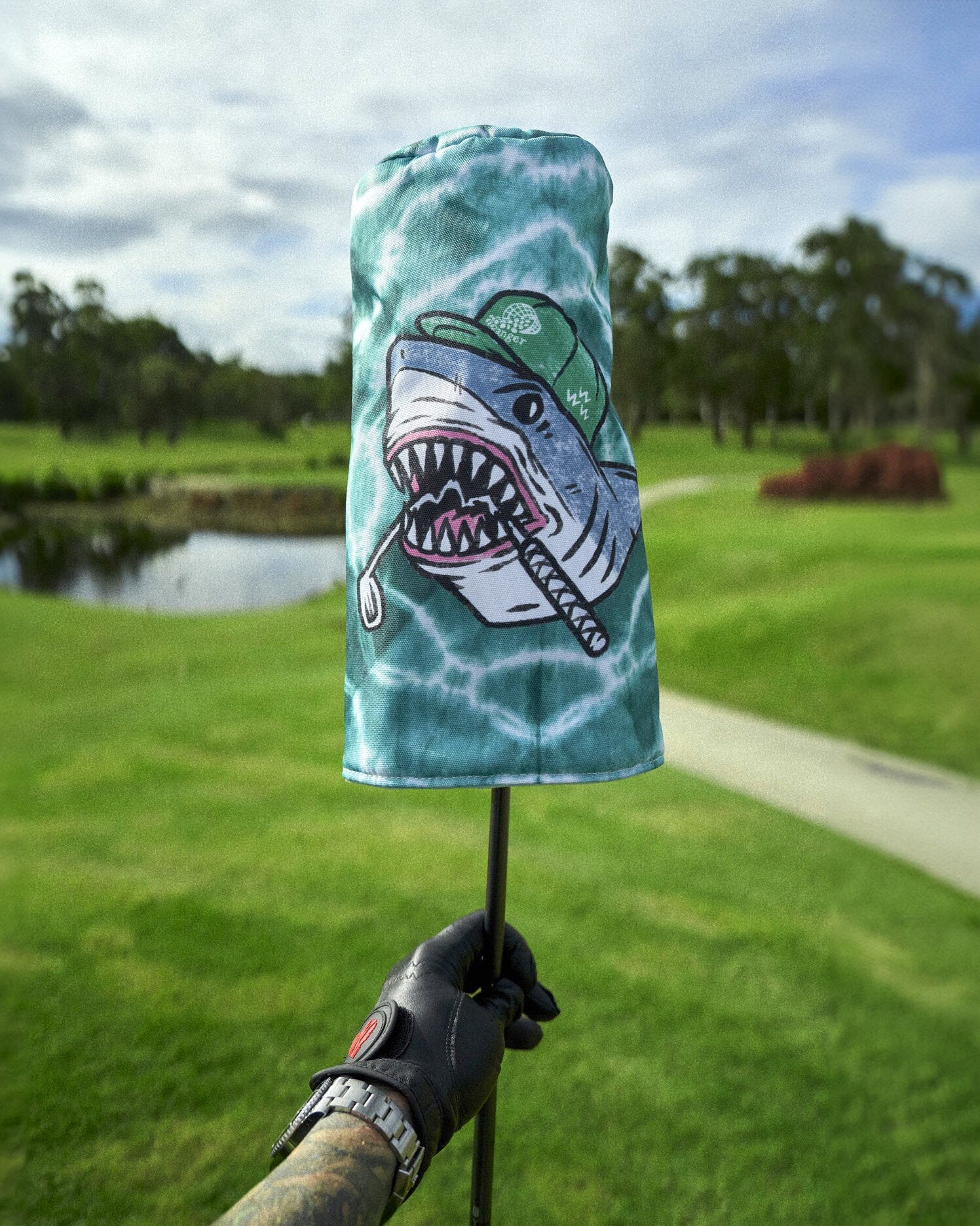 A tiedye headcover featuring a shark graphic wearing a green cap, held above a golf course.