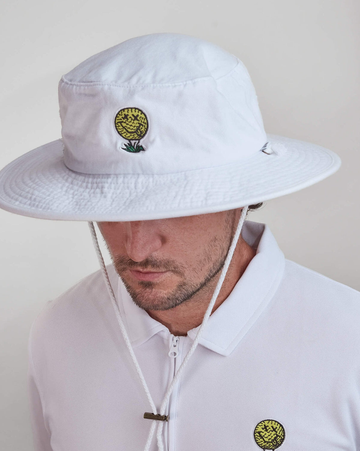 A man wearing a white cotton boonie hat with a small smiley face golf ball design on the front.