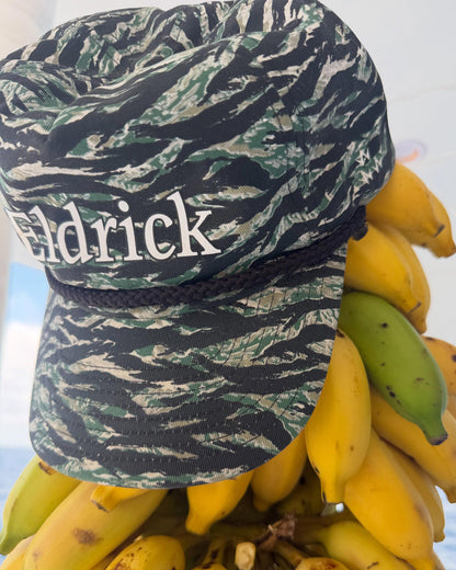 A close-up of a baseball cap with a tiger-stripe camouflage pattern in green, black, and beige, featuring the name 'Eldrick' in white text on the front. The cap is styled on top of a bunch of yellow and green bananas against a bright, overexposed background.