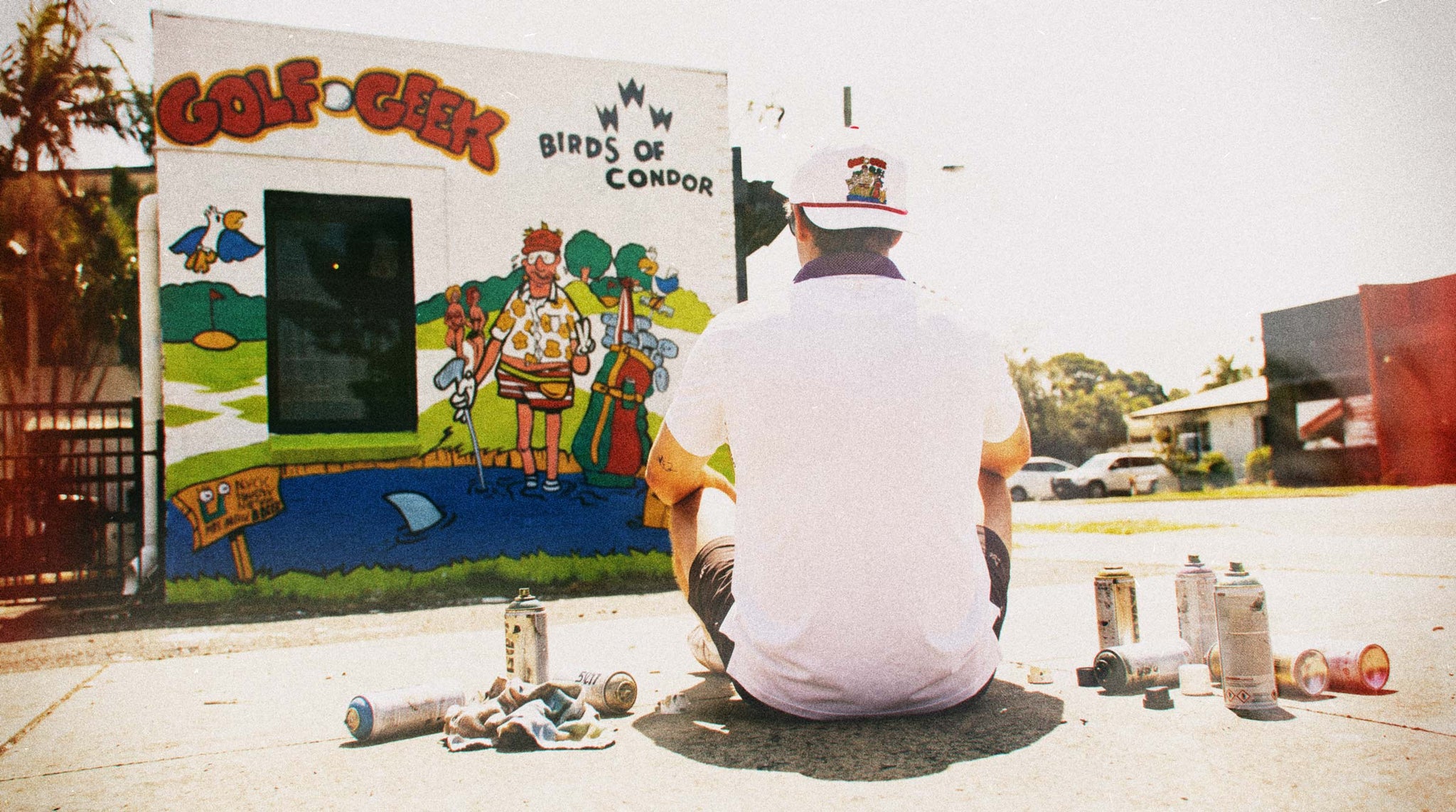 A man sits on the ground facing a colorful painted wall mural that reads 'GOLF GEEK' and 'BIRDS OF CONDOR,' with spray paint cans scattered around him in an outdoor street setting.