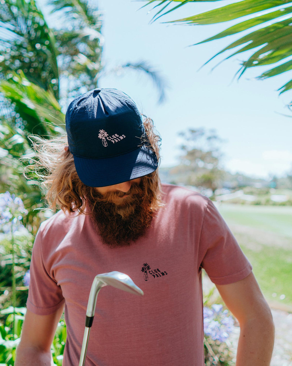 A man wearing a navy snapback with pale pink palm print graphic and a curved brim outdoors.